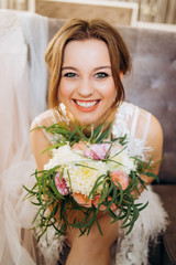 A smiling bride with a bouquet in her hands sits on a soft chair in the room.