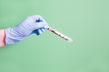 one hundred dollar bill in a glass test tube, a hand in a blue rubber disposable medical glove holds a test tube on a green background, copy space, crisis against the backdrop of the pandemic 2020