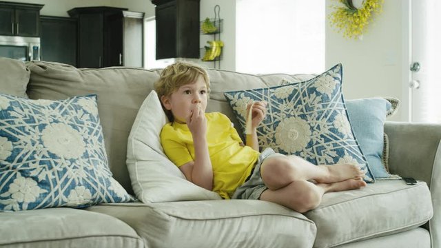 4 year old boy lounges on couch eating string cheese while watching television
