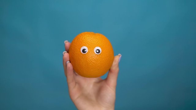 Orange with eyes in a woman hand. Orange jumps into the frame and looks around on a blue background. Woman hand close-up.