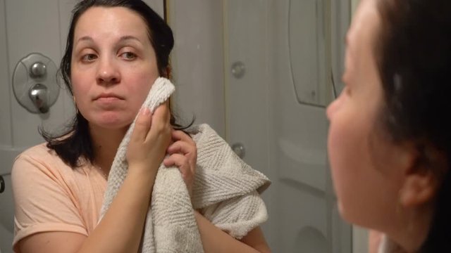 A young woman is standing in the bathroom. He wipes his face with a towel. Reflection of a man in the mirror. Hygienic procedure. Half-length portrait.