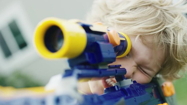 Blonde boy looks through scope of toy sniper rifle and smilles before firing