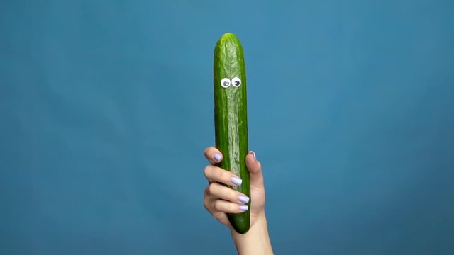 Cucumber with eyes in a woman hand close-up. Cucumber shakes and twists eyes on a blue background. Slow motion
