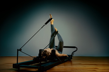 Young girl doing pilates exercises with a reformer bed. Beautiful slim fitness trainer on reformer gray background, low key, art light. Fitness concept