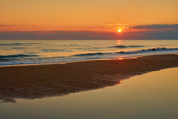 Colorful sunset at the tropical beach, sun behind clouds reflects on water and waves with foam hitting sand.