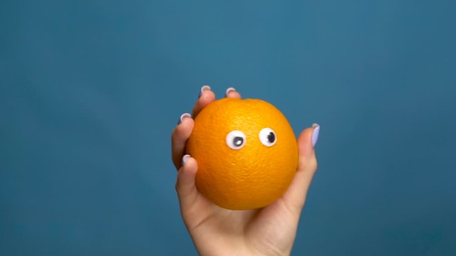 Orange with eyes in a woman hand close-up. Orange shakes and twists eyes on a blue background. Slow motion.