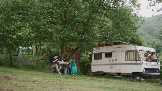Young couple relaxing on picnic under big tree near trailer. Family vacation