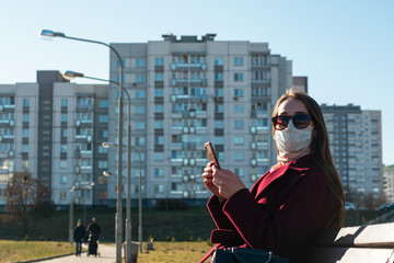 girl in sunglasses sitting on a bench and looking at a cell phone. On the face a protective mask...