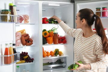 Young woman taking cucumber out of refrigerator indoors