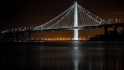 Fototapeta premium Bay Bridge at night from Treasure Island. Port of Oakland in the background. Long exposure with water reflection.