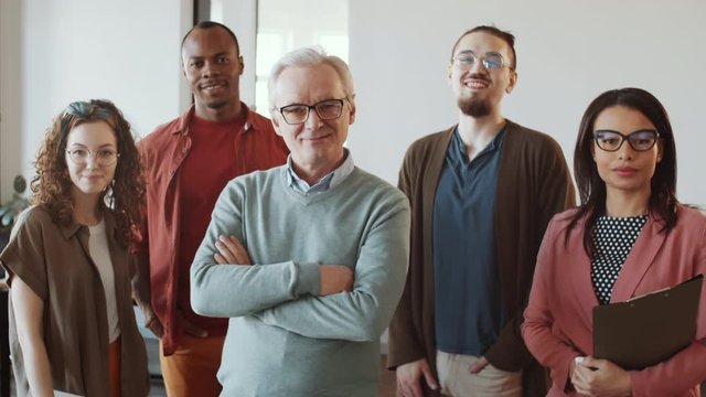 Cheerful senior manager and young multiethnic male and female employees in casual clothes standing together in office, smiling and posing for camera during workday