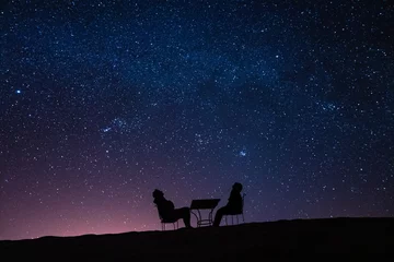 Tuinposter young couple sitting at a table on a desert dune while talking, relaxing and observing the stars and the milky way above them © ylenia cancelli