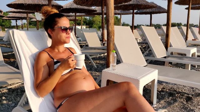 Beautiful pregnant woman in swimsuit and sunglasses drinking tea on the beach while looking at the sunrise