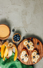 Buckwheat healthy bread with peanut butter, banana and blueberry on wooden board over concrete background. Top view. Flat lay. Summer breakfast.