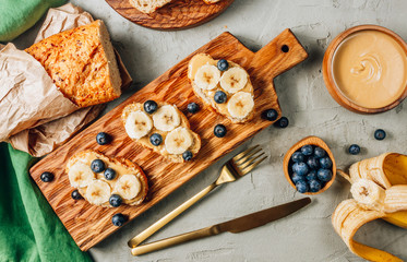 Buckwheat healthy bread with peanut butter, banana and blueberry on wooden board over concrete background. Top view. Flat lay. Summer breakfast.