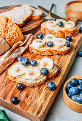 Buckwheat healthy bread with peanut butter, banana and blueberry on wooden board over concrete background. Selective focus. Summer breakfast.