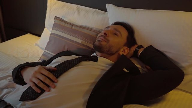 happy businessman in suit resting on bed in hotel room
