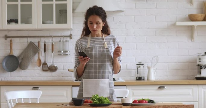Happy young woman preparing vegetable meal searching checking online vegan recipe in smartphone, smiling vegetarian girl cooking healthy food salad using mobile cookbook menu apps in modern kitchen