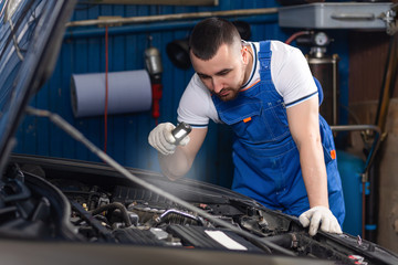 Handsome young male auto mechanic in special uniform clothes holding a flashlight, looking for...