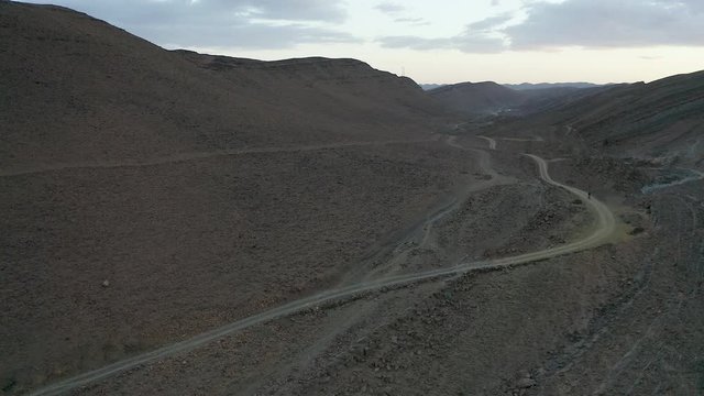 An aerial boom down and push-in on a solitary cyclist or bicycle rider, bikepacking uphill through a colorful but barren, rocky, desolate desert landscape at dusk or dawn.