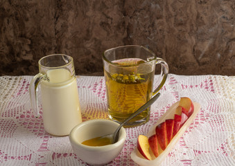 Peppermint tea, with floating mint leaves, in a clear glass cup, a jug with cow's milk, red apple cut into pieces to accompany the tea and a ceramic pot with honey to sweeten the tea. With wooden back