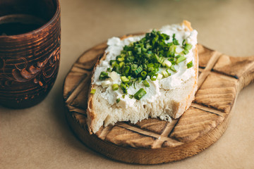 Breakfast on a craft background, a ceramic mug with coffee and a sandwich with curd cheese and green onions on a wooden stand; warm tinting, we cook at home with pleasure, a recipe for spring mood