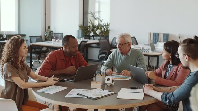 Senior male CEO showing business plan on paper and explaining it to young multiethnic team while holding meeting in office