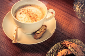 Eine Kaffeetasse mit gerösteten Kaffeebohnen auf einem hölzernenen Tisch. Sonneneinstrahlung, Reflexionen. Ein Cappuccino mit Milchschaum und Keksen. Serviert in einem Café. Milchkaffee