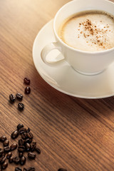 Eine Kaffeetasse mit gerösteten Kaffeebohnen auf einem hölzernenen Tisch. Sonneneinstrahlung, Reflexionen. Ein Cappuccino mit Milchschaum und gemahlenem Kaffee. Serviert in einem Café. Milchkaffee