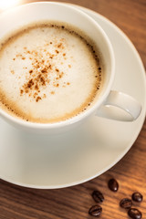 Eine Kaffeetasse mit gerösteten Kaffeebohnen auf einem hölzernenen Tisch. Sonneneinstrahlung, Reflexionen. Ein Cappuccino mit Milchschaum und gemahlenem Kaffee. Serviert in einem Café. Milchkaffee