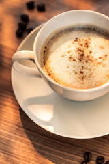 Eine Kaffeetasse mit gerösteten Kaffeebohnen auf einem hölzernenen Tisch. Sonneneinstrahlung, Reflexionen. Ein Cappuccino mit Milchschaum und gemahlenem Kaffee. Serviert in einem Café. Milchkaffee