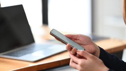 Cropped image of businesswoman's hand holding a smartphone over computer laptop putting on wooden working desk as background. Technology and lifestyle concept.