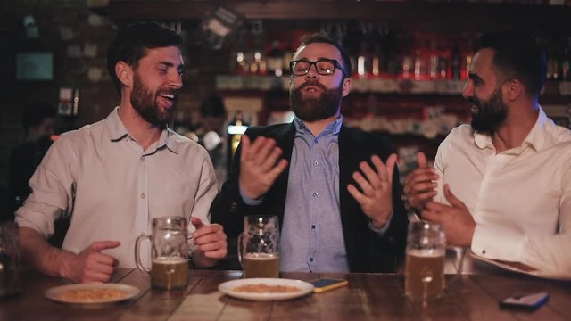 Two cheerful men listening to his bearded friend funny story and laughing while sitting in pub. Male friends resting after work day, joking and having good time together.