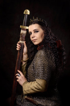 A dark-haired woman with a sword and crown, a Scandinavian warrior in a chain mail against a dark background