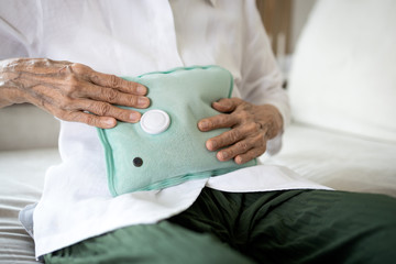 Close up of senior woman belly with hot water bag sitting on the couch at home,sick elderly people...