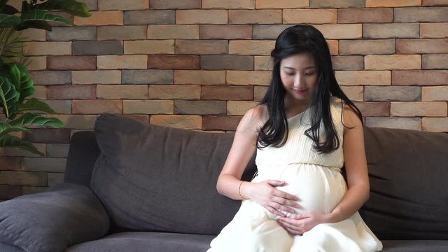 Close-up of young Asian pregnant maternity woman touching and holding an unborn child inside tummy sits on sofa at home