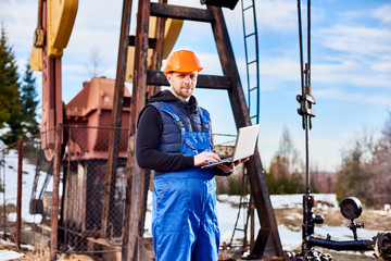 Portrait of petrolium engineer, wearing blue overalls and orange helmet, with a laptop, standing...