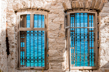 Two windows with rusty wrought iron fences inside light brown brick house walls, turquoise see water surface with ribbles