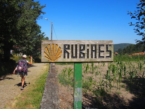 Pilgrim walking with wooden signboard with the inscription "RUBiAES" and yellow scallop shell mark on the road to Santiago de Compostela, Camino de Santiago, Way of St. James, Ponte de Lima to Rubiaes