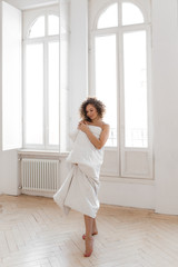 Beautiful sweet cheerful tender happy natural sleepy girl with Afro curls wrapped in a blanket wakes up and enjoys the morning looks out the window against the background of large vintage Windows
