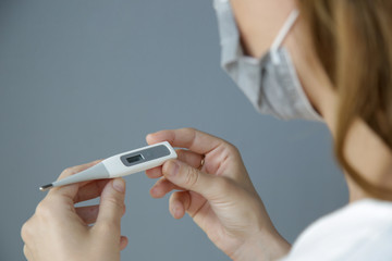 Close up portrait of young woman wearing medical mask and holding thermometer. Fever and symptoms of virus infection. COVID 19 novel Coronavirus outbreak concept image.