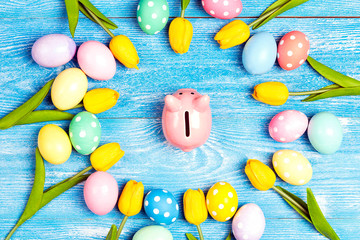 Piggy bank surrounded by Easter eggs and tulips on a blue wooden background.