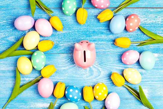 Piggy bank surrounded by Easter eggs and tulips on a blue wooden background.