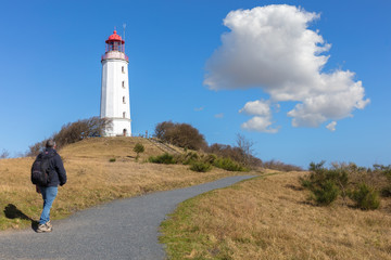 Fototapeta na wymiar The Dornbusch lighthouse on the German island of Hiddensee in the Baltic Sea. It is a beautiful winter day with blue sky and clouds. In the foreground a hiker with a backpack.
