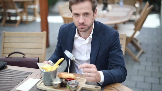 young smiling businessman is eating his hamburger from his plate of  French fries, sauces, sitting at a table with his briefcase next to him at a terrace