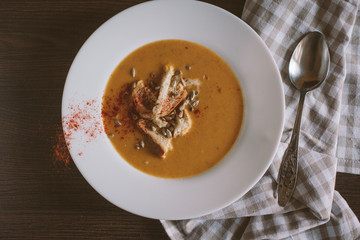 Pumpkin puree soup with crackers and seeds sprinkled with chili peppers in a large plate on a dark wooden background. Next to a spoon and a linen towel. Healthy and wholesome food.