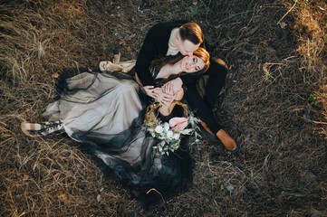 Loving groom in a black cardigan kisses a cute bride in an expensive dress, sitting in the woods in nature. Wedding portrait of smiling newlyweds. Photography, concept.