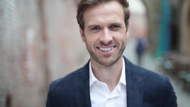 close up of a young sexy businessman wearing a navy suit and looking at camera while smiling wide, outdoor