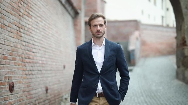 smart casual businessman wearing a navy suit is walking with one hand in pocket and looking around near a brick wall