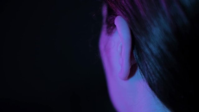 Close up shot on womans ear and hand putting in and out earphone; purple and blue light, girl wearing headphones and listening to music, handheld camera